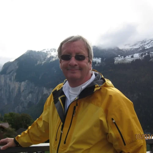 Jim in a yellow rain jacket standing before snow-capped mountains and dramatic alpine valley scenery.