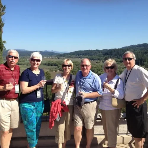 Jim stands with his wife and 4 other adults smiling and holding wine glasses outdoors at a vineyard with rolling hills and greenery in the background.