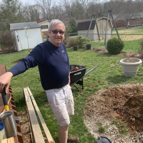 Jim in navy shirt and white shorts smiling outdoors near lumber and a wheelbarrow in a backyard garden.