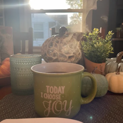 A coffee cup that says "today I choose joy" sits on a dining room table surrounded by candles, plants, and decorative center piece elements