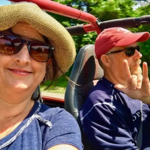 Carrie and her husband riding in a jeep. Carrie's husband signs "I love you" with his left hand.