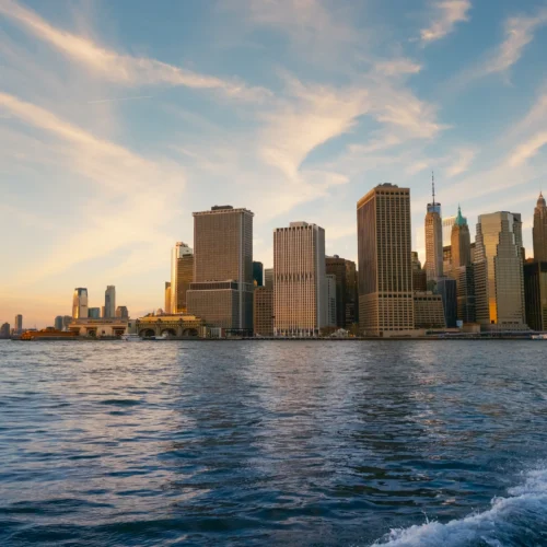 Manhattan skyline at golden sunset, viewed from water with rippling waves in foreground and dramatic cloudy sky.