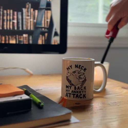 Caitlynn lighting a candle on her desk. On the monitor, ambient library and thunder storm sounds play. On the desk, there is a coffee mug that says "my neck, my back, my anxiety attack" alongside sketchbooks, markers, and post-it notes
