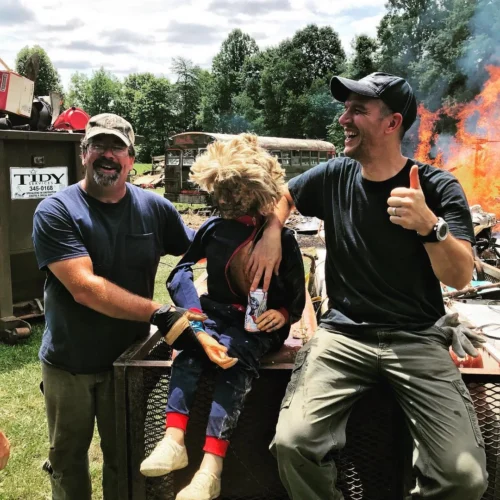 Two laughing men support a well-worn CPR dummy while a large bonfire burns in the background near old school buses