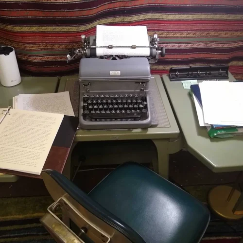 A vintage gray Royal typewriter with paper loaded, sitting on an industrial green desk between a three-ring binder of typed pages and stacks of paper, with a striped woven wall hanging behind and a forest green mid-century office chair in the foreground.