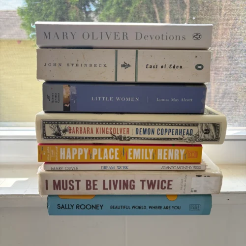 A stack of Emily's favorite books, including Devotions and Dream Work by Mary Oliver, East of Eden by John Steinbeck, Little Women by Louisa may Aleaott, Demon Copperhead by Barbara Kingsolver, Happy Place by Emily Henry, I must be living twice by Eileen Myles, and Beautiful World Where Are You by Sally Rooney