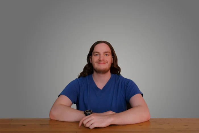 Carter Igo seated at a wooden table, wearing a blue v-neck shirt, arms crossed, smiling against a gray background