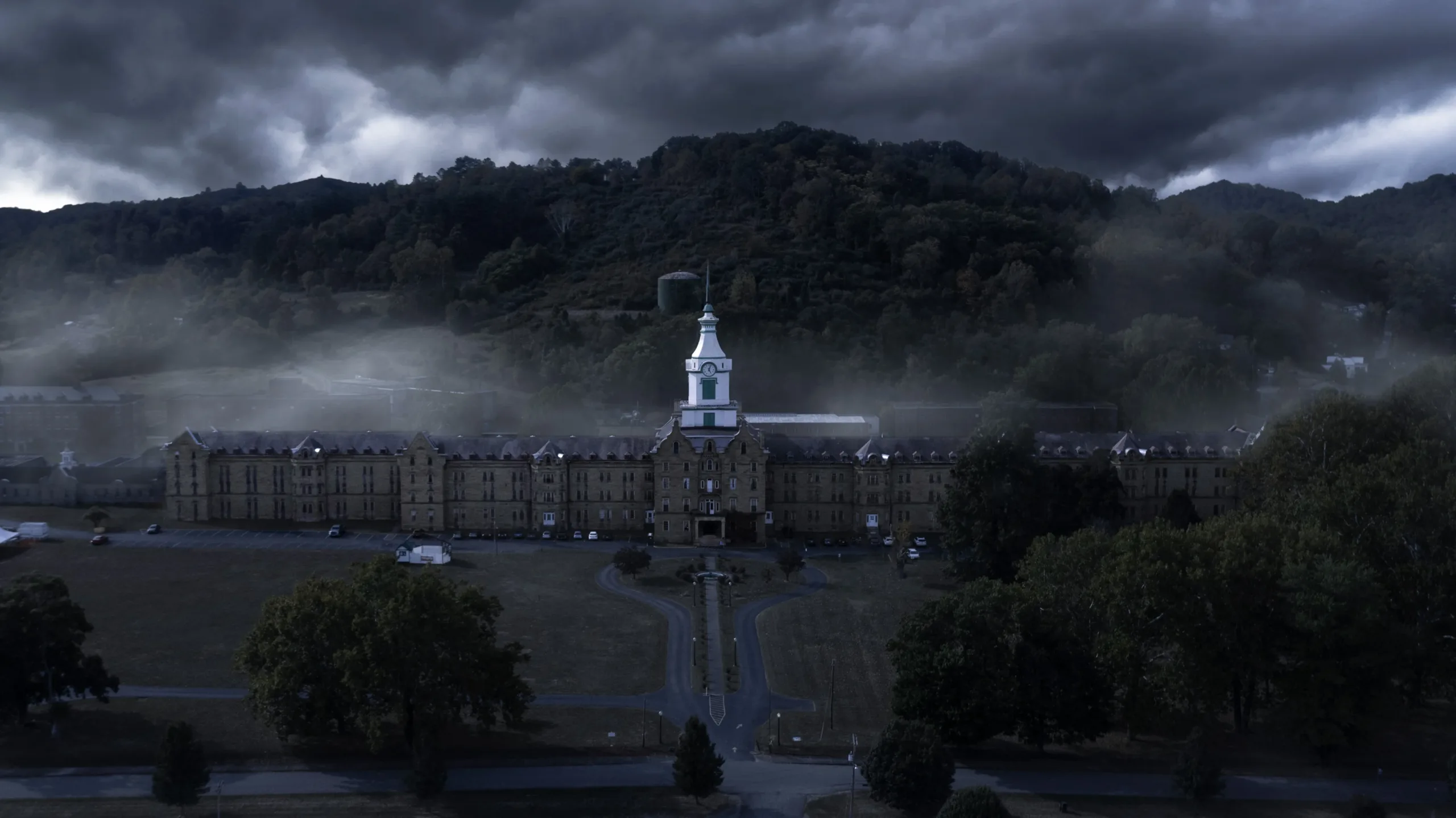 A cinematic aerial image of the Trans-Allegheny Asylum at Weston, West Virginia. The sky is dark and stormy, and fog sits heavily around the building.