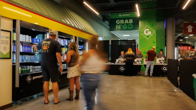 A group of people navigate the GoMart Grab N' Go register area in the Charleston Civic Center. Multiple people browse the drink selection in the branded coolers. Prominent Signage is featured, indicating the purpose of the area. The wall behind the register area features colorful, illustrated graphics - reflective of the GoMart brand