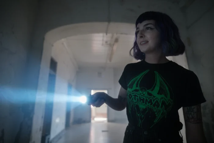 A cinematic image of a paranormal explorer at one of the many Paranormal Trail locations. She is shining a flashlight down a dark hallway, and is wearing a t-shirt featuring the local cryptid, Mothman