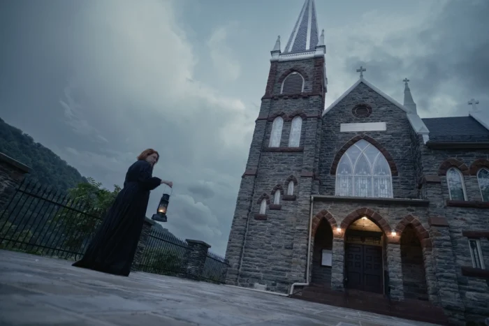 A cinematic image of the Harper's Ferry Ghost Tour. The tour guide is shown from a low angle in front of a gothic revival style chapel. The tour guide is wearing an all-black, period appropriate dress and holding a lit lantern. The Appalachian mountains are visible in the background beneath an overcast and eerie sky.