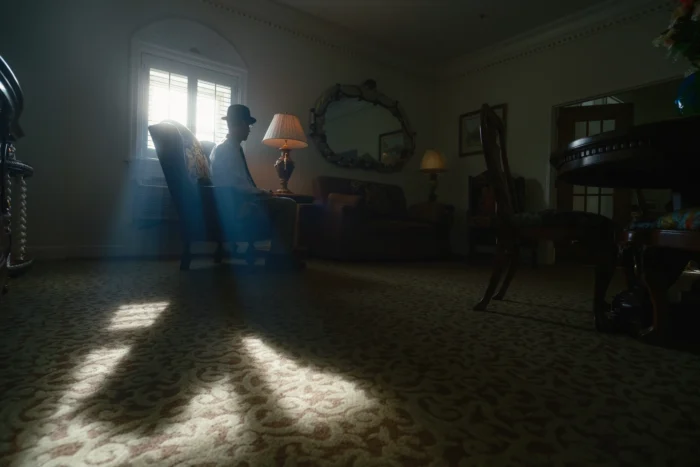 A cinematic image of a man in a bowler hat, sitting alone in front of a window in the Blennerhassett Hotel. The room is dimly lit, and the window casts a long and spectral shadow of the chair across the floor. The seated man's shadow is absent.