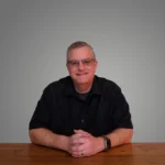 Aaron Gooden sitting at a wooden table, wearing glasses and a black shirt, smiling against a gray background.