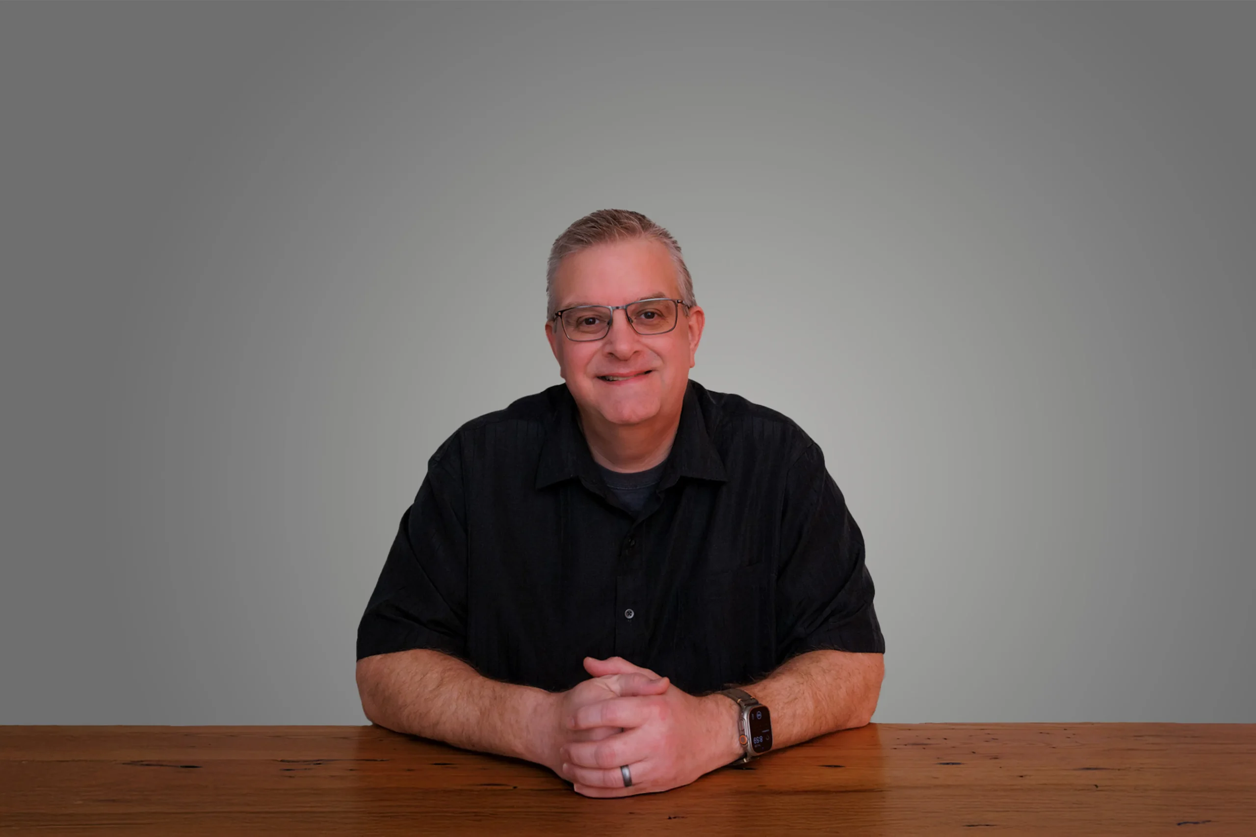 Aaron Gooden sitting at a wooden table, wearing glasses and a black shirt, smiling against a gray background.