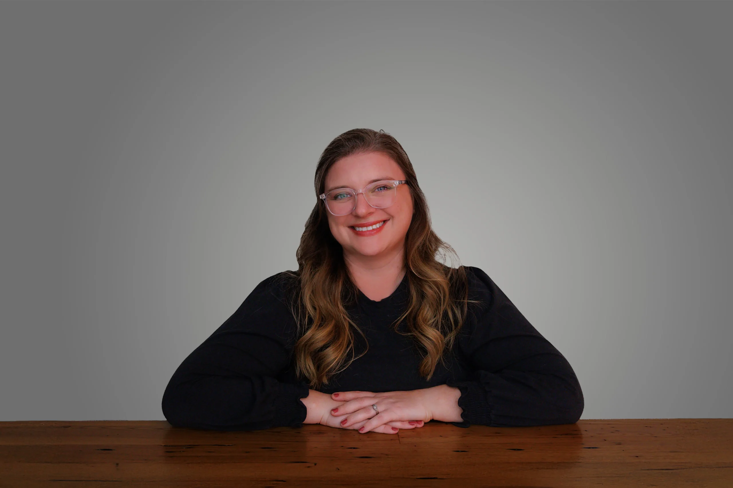 Alaina Moore smiling, sitting at a wooden table against a gray background.