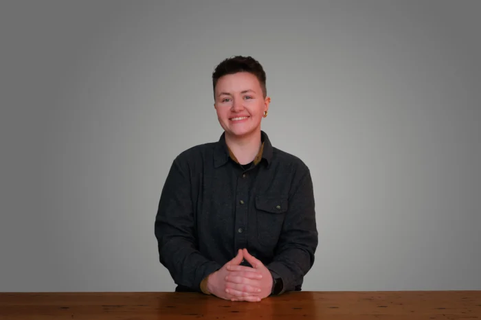 Caitlynn Jones seated at a wooden table, wearing a dark gray button-up shirt, hands folded, smiling against a gray background