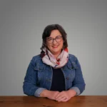 Carrie Nesselrode smiling, sitting at a wooden table, wearing a denim jacket and floral scarf.