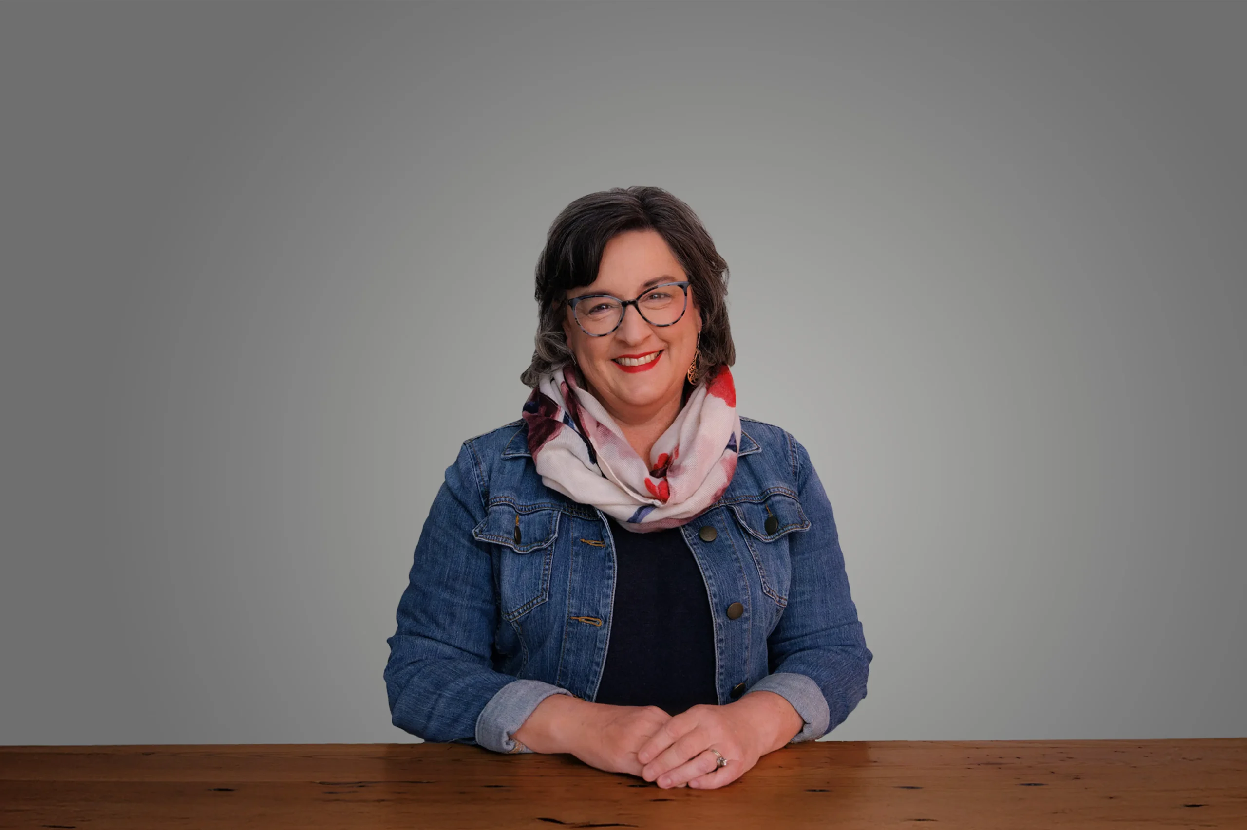 Carrie Nesselrode smiling, sitting at a wooden table, wearing a denim jacket and floral scarf.