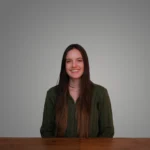 Emily Akers smiling, sitting at a wooden table against a gray background.