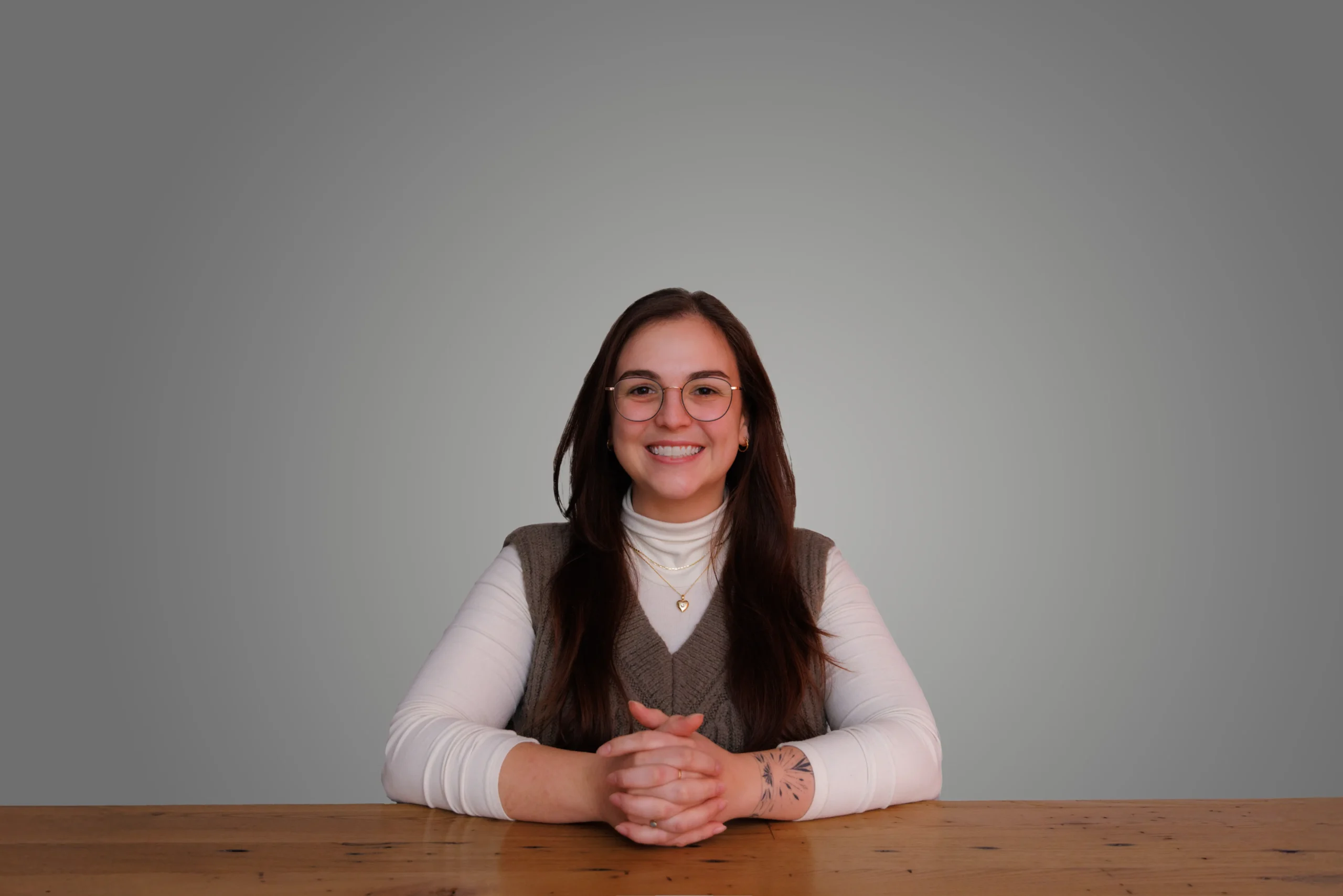 Emma Malinoski smiling, seated at a wooden table with a gray background, wearing glasses, a white shirt, and a gray vest.