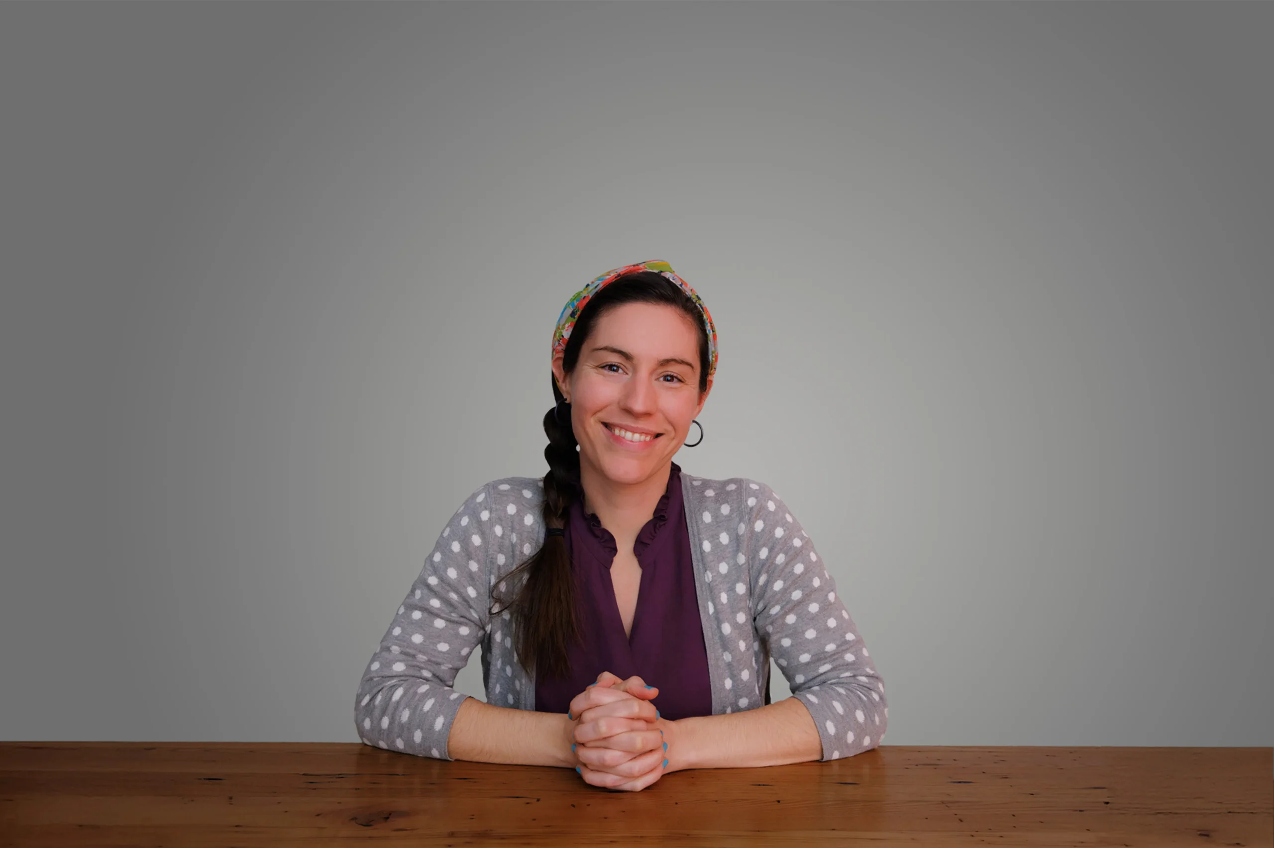 Hilary Roush Freeman smiling, seated at a wooden table with hands clasped, wearing a polka dot sweater and a colorful headband.