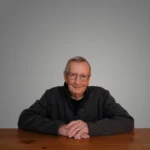 Jim Strader sitting at a wooden table against a plain gray background, wearing glasses and a dark sweater.
