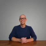 Jonathan Danz smiling and sitting at a wooden table against a plain background.