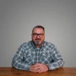 Pat Strader wearing glasses and a plaid shirt, sitting at a wooden table against a gray background.