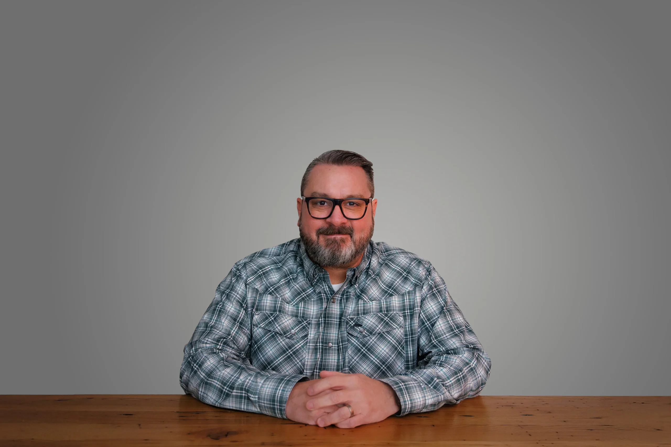 Pat Strader wearing glasses and a plaid shirt, sitting at a wooden table against a gray background.