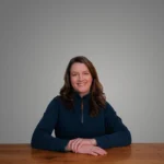 Sarah Powell smiling, sitting at a wooden table against a plain background.