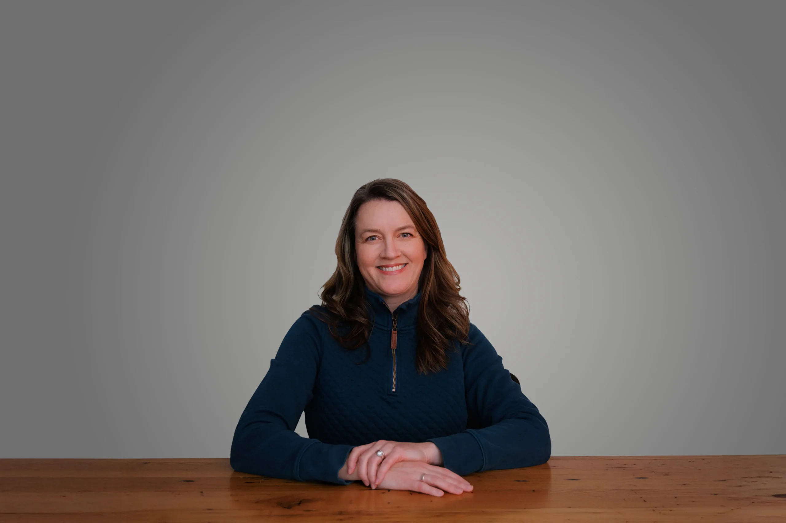 Sarah Powell smiling, sitting at a wooden table against a plain background.