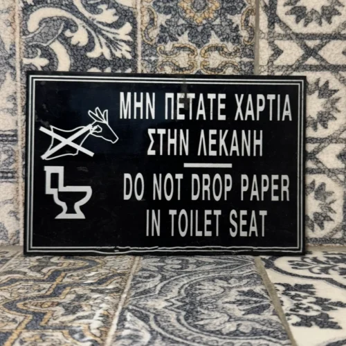 A tiled wall and a sign in greek and english reminding bathroom users to not put paper down the toilet