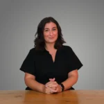 Vasilia Scouras in a black shirt sits at a wooden table, hands clasped, with a neutral background.