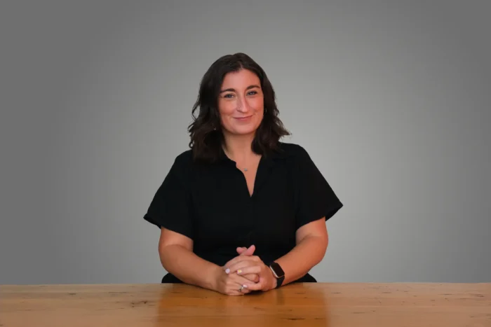 Vasilia Scouras in a black shirt sits at a wooden table, hands clasped, with a neutral background.