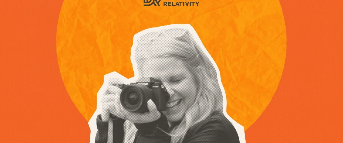 Woman photographing with a camera, smiling, against an orange circular background. Digital Relativity logo in top left corner.