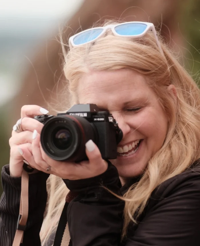 Abbey Reifsnyder with sunglasses on head, taking a photo with a Fujifilm camera in an outdoor setting.