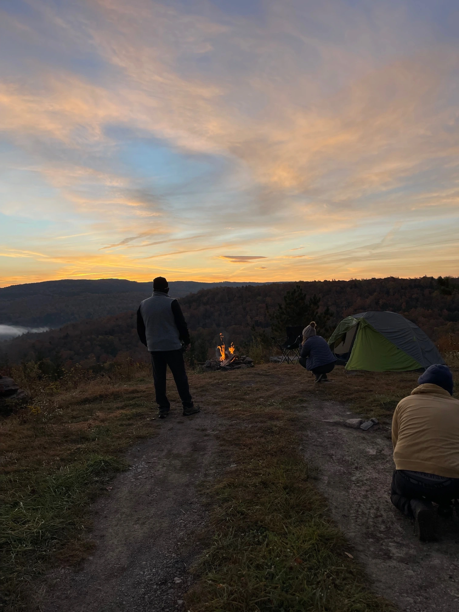 People camping on a hilltop at sunset, with a tent and campfire under a colorful sky.