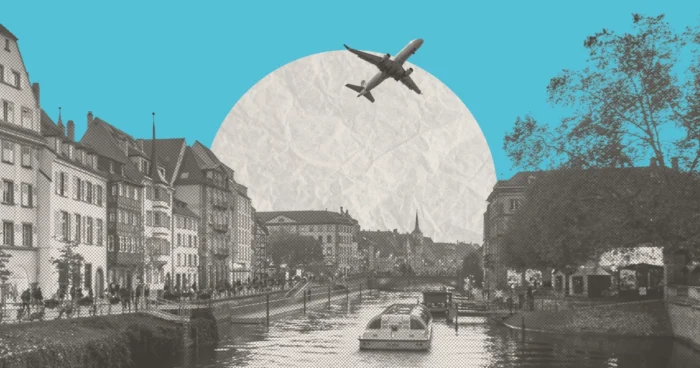 Black and white cityscape of a canal with historic buildings, a boat, and a plane flying overhead against a teal sky.