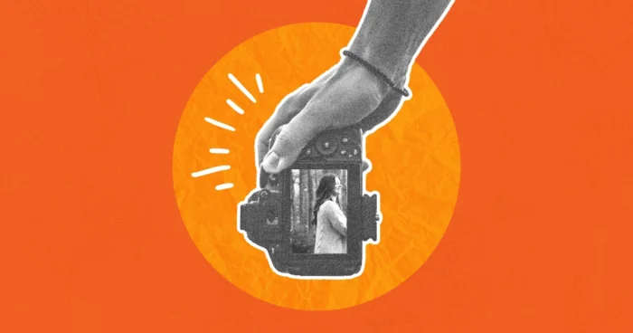Hand holding a vintage camera with a small figure visible inside the lens, on an orange background.