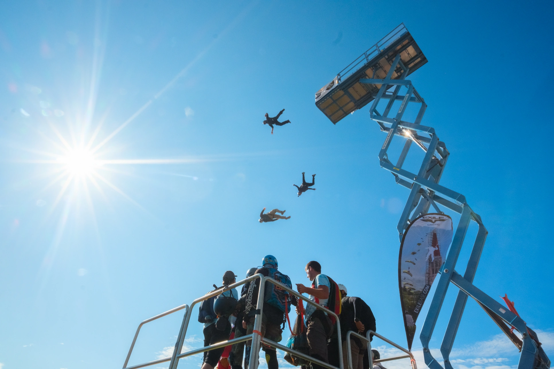 People on an elevated platform watch aircraft flying overhead against a bright blue sky with a sunburst.
