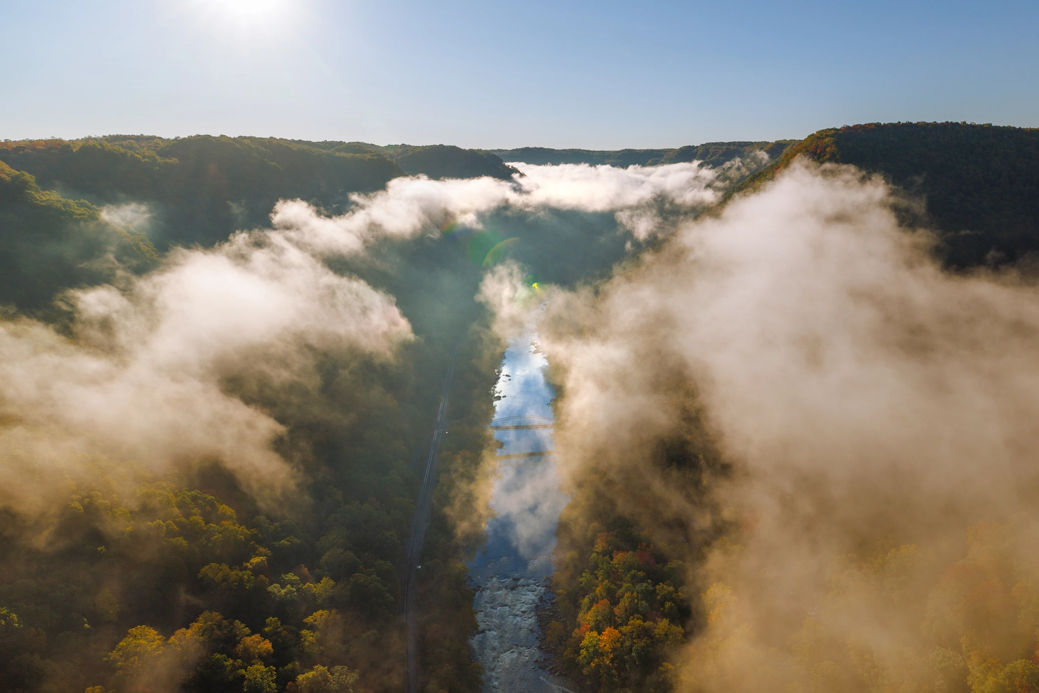 Aerial view of a misty river valley with fall foliage, low clouds, and a winding river under a clear blue sky.