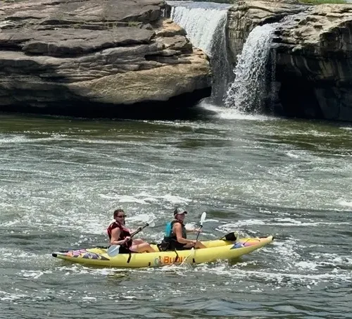 Hannah and her friend Sarah on a bright yellow tandem kayak with a waterfall visible in the background